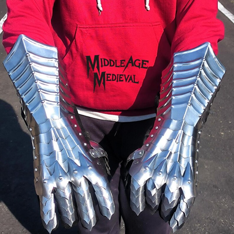 May include: A pair of shiny, silver metal gauntlets, designed with articulated plates for flexibility. The gauntlets have a medieval aesthetic, with pointed edges and rivets. The text "Middle Age Medieval" is visible on a red hoodie.