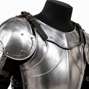May include: A polished steel breastplate and pauldrons set featuring curved metal plates, rounded shoulder guards, and multiple silver-toned rivets. The armor is secured with black leather straps and brass-colored metal buckles over a black base.