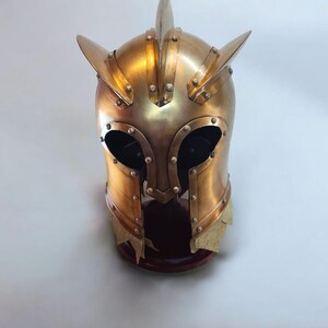 Op de afbeelding: Een goudkleurige middeleeuwse helm met een puntige top en zijaccenten. De helm heeft oogopeningen en is geniet. De helm wordt tentoongesteld op een donkere houten standaard.