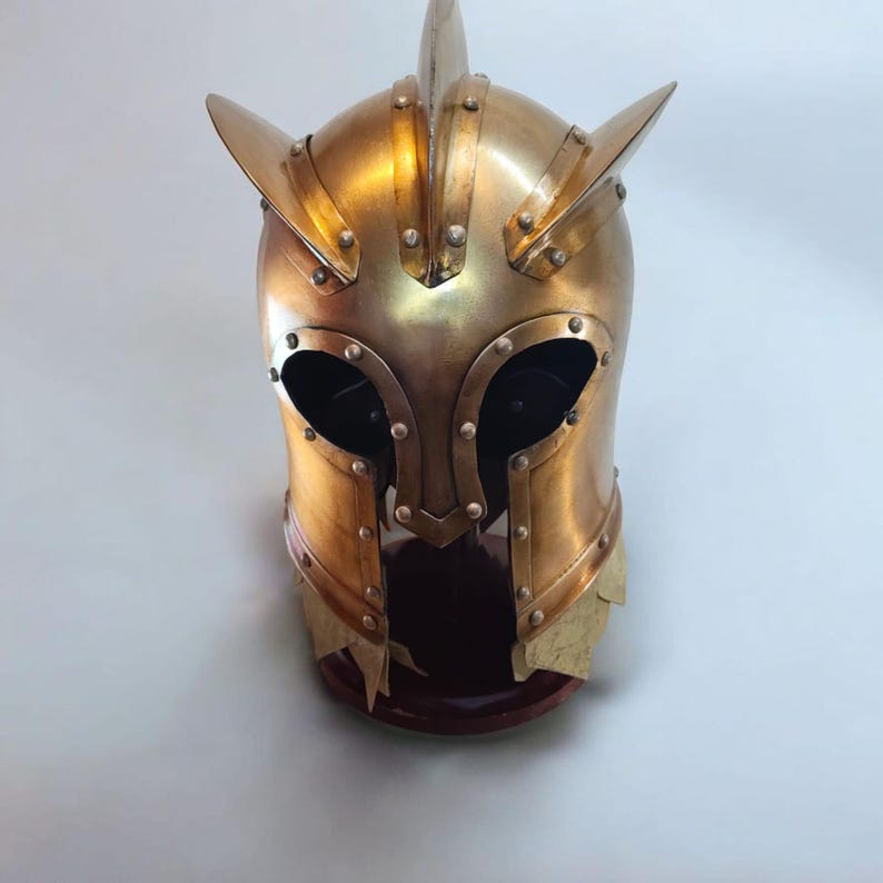 Op de afbeelding: Een goudkleurige middeleeuwse helm met een puntige top en zijaccenten. De helm heeft oogopeningen en is geniet. De helm wordt tentoongesteld op een donkere houten standaard.