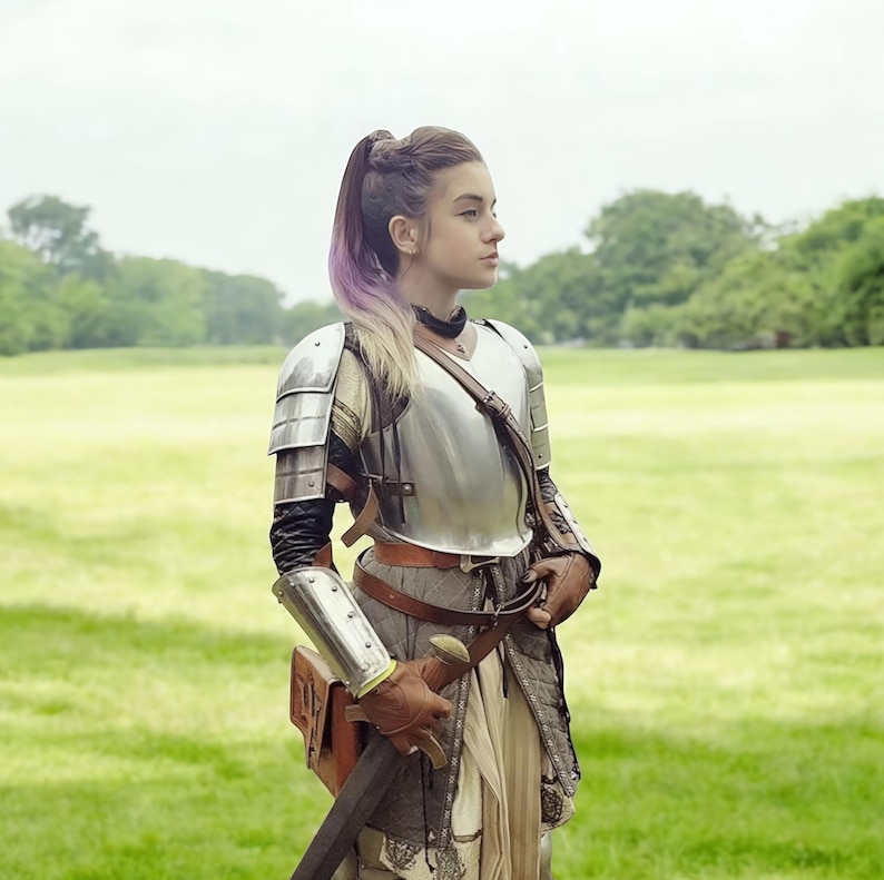 May include: A person in medieval armour, including a silver breastplate, shoulder guards, and arm protection. They are holding a sword and wearing brown leather gloves and a belt. The background is a green field and trees, with a cloudy sky overhead.