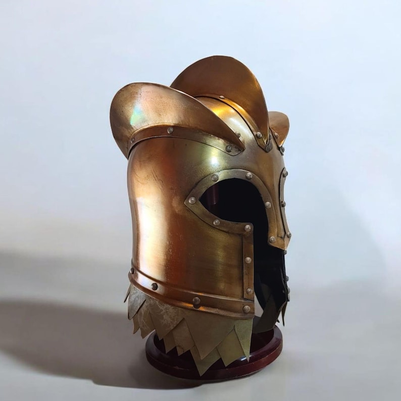 Op de afbeelding: Een bronzen helm met een afgeronde bovenkant en een vizieropening. De helm heeft een decoratief kam en is langs de randen geklonken. Het onderste deel heeft overlappende metalen platen. Het wordt tentoongesteld op een houten standaard.