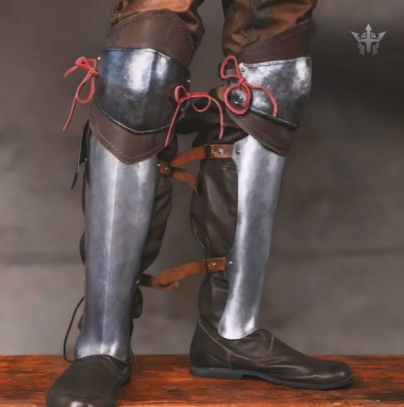 May include: A pair of brown leather boots featuring metallic greaves and knee guards. The armour plates are secured with brown leather straps and buckles, while the knee sections are fastened with thin red cord ties. A small silver logo is visible.