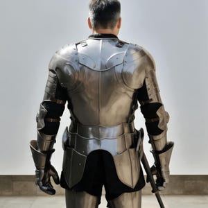 May include: A polished steel suit of armor featuring a breastplate, pauldrons, articulated arm guards, and gauntlets. The metallic plates are secured with brown leather straps over a black garment, covering the torso, shoulders, and arms.