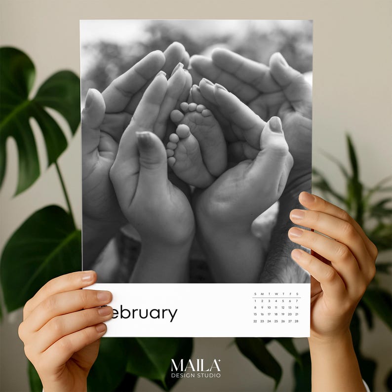 May include: Monochrome photograph of a calendar page showing a baby's feet held by adult hands. The word "february" is printed at the bottom, along with a small calendar grid. The "MAILA DESIGN STUDIO" logo is also visible.