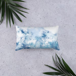 May include: A rectangular lumbar cushion with a blue and white watercolour design of clouds. The cushion is on a grey concrete background.