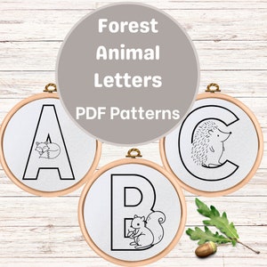 May include: Three embroidery hoops with black and white illustrations of the letters A, B, and C. Each letter has a different forest animal inside: a fox, a squirrel, and a hedgehog. The hoops are on a white wood background. The text "Forest Animal Letters PDF Patterns" is above the hoops.