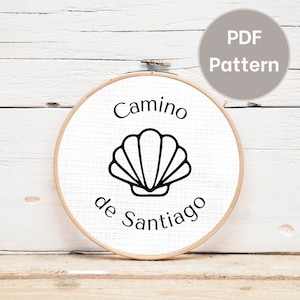 May include: A white embroidery hoop with a black outline of a scallop shell and the text "Camino de Santiago".