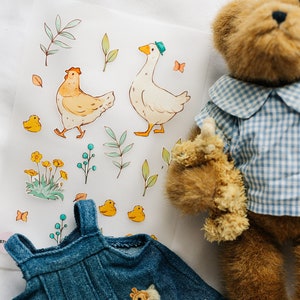 Peut inclure: Une robe en jean bleu avec une applique de girafe, un ours en peluche portant une chemise à carreaux bleu et blanc, et une feuille d'autocollants avec une poule, une oie et des fleurs.