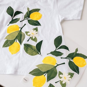 Könnte beinhalten: Weißes T-Shirt mit Bügelbildern von Zitronen und Blumen. Die Aufkleber sind grün, gelb und weiß.