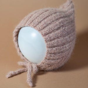 Pode incluir: Um gorro de bebê de malha canelada rosa claro, com uma parte superior pontiaguda e laços. O gorro é feito de um fio macio e texturizado e é exibido em uma forma arredondada branca sobre um fundo neutro. O design do gorro é simples e clássico.