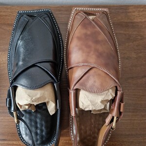 May include: A pair of Peshawari sandals, one black and one brown, displayed on a wooden surface. The sandals feature a distinctive design with a strap and buckle. The black sandal has a black strap, and the brown sandal has a brown strap.