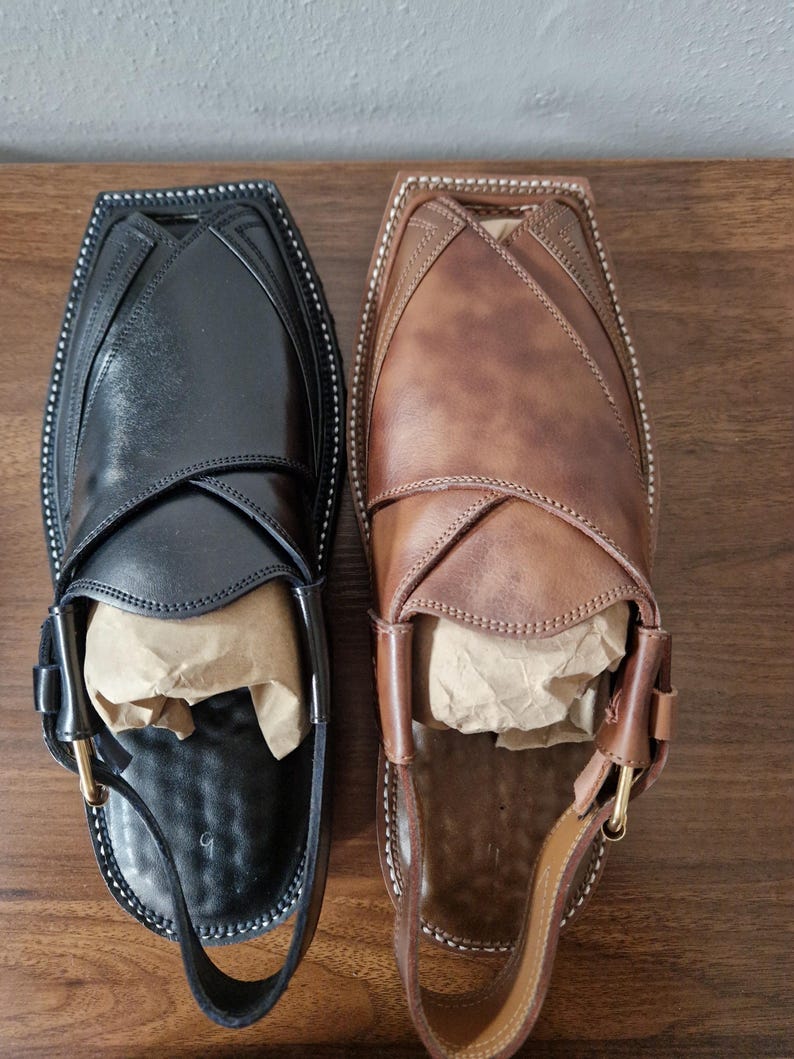May include: A pair of Peshawari sandals, one black and one brown, displayed on a wooden surface. The sandals feature a distinctive design with a strap and buckle. The black sandal has a black strap, and the brown sandal has a brown strap.