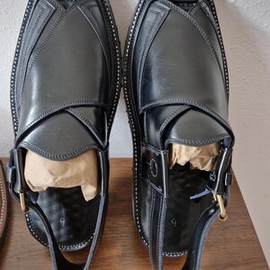May include: A pair of black leather Peshawari sandals. These traditional sandals have a square toe, detailed stitching, and a buckle fastening. The sandals are presented on a wooden surface.