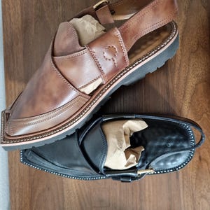 May include: Two leather sandals, one brown and one black, are displayed on a wooden surface. The brown sandal has a strap with a buckle and decorative stitching. The black sandal also has a buckle and a similar design.