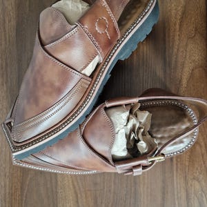 May include: A pair of brown leather sandals with a slingback design. The sandals feature a stitched border, a black sole, and a buckle fastening. The leather has a distressed look. The sandals are displayed on a wooden surface.