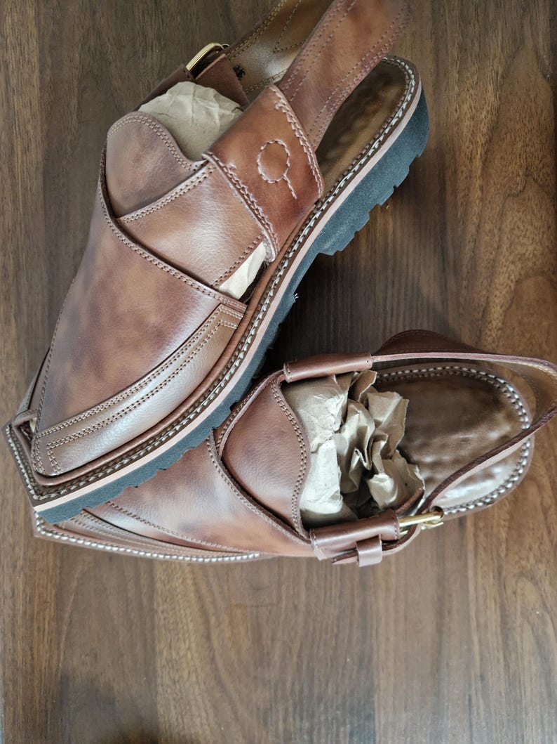 May include: A pair of brown leather sandals with a slingback design. The sandals feature a stitched border, a black sole, and a buckle fastening. The leather has a distressed look. The sandals are displayed on a wooden surface.
