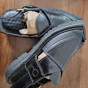 May include: Black leather sandals with a thick sole and buckle fastening. The sandals feature an ankle strap and a strap across the foot. White stitching details the design. The sandals are new.