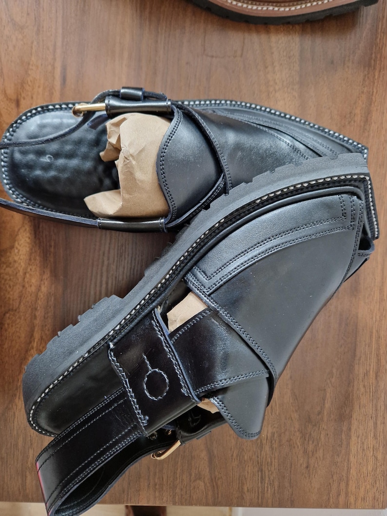 May include: Black leather sandals with a thick sole and buckle fastening. The sandals feature an ankle strap and a strap across the foot. White stitching details the design. The sandals are new.