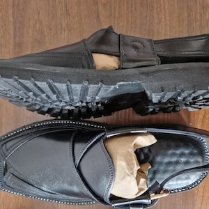 May include: Two black leather sandals with chunky, ridged soles. One sandal is upside down, showing the sole's pattern. The other sandal has a strap and buckle fastening. Both sandals have a polished finish and are on a wooden surface.