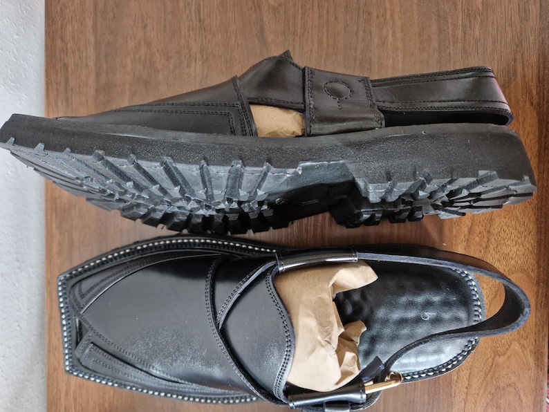 May include: Two black leather sandals with chunky, ridged soles. One sandal is upside down, showing the sole's pattern. The other sandal has a strap and buckle fastening. Both sandals have a polished finish and are on a wooden surface.