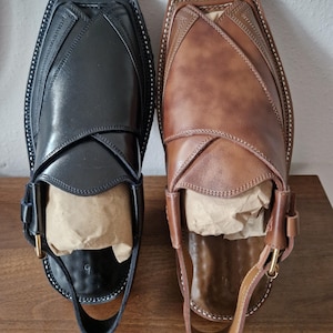 May include: Two leather Peshawari sandals, in black and brown, are shown. The sandals have a distinctive design with a strap and buckle. The soles are stitched with white thread. The sandals are filled with paper for shape.