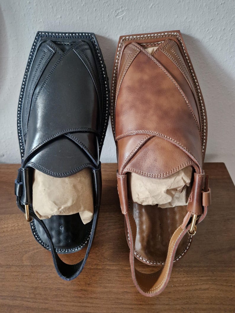 May include: Two leather Peshawari sandals, in black and brown, are shown. The sandals have a distinctive design with a strap and buckle. The soles are stitched with white thread. The sandals are filled with paper for shape.