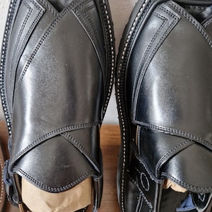 May include: Pair of black leather Peshawari sandals. The sandals feature a square toe design with crisscross straps and decorative stitching. The soles are black with white stitching around the edges. The sandals are new and unworn.