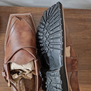 May include: A pair of brown leather sandals with a distinctive design. The sandals have a square toe, detailed stitching, and a strap with a buckle. The black soles have a rugged tread pattern. The sandals are new and filled with paper.