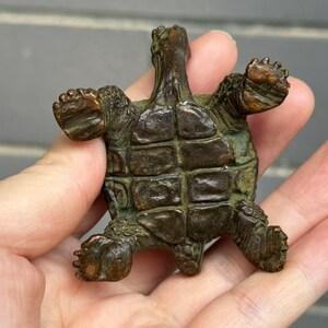 May include: A bronze turtle figurine with outstretched limbs. The turtle has a detailed shell and textured skin, with a weathered, aged appearance. The figurine is held in a hand, showcasing its size and intricate design.