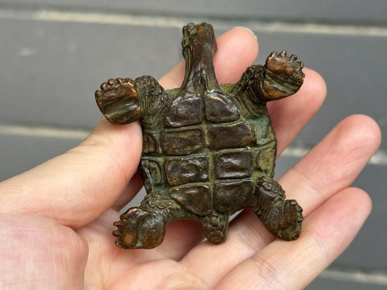 May include: A bronze turtle figurine with outstretched limbs. The turtle has a detailed shell and textured skin, with a weathered, aged appearance. The figurine is held in a hand, showcasing its size and intricate design.