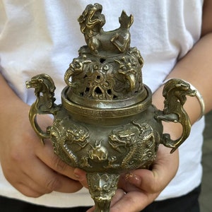 May include: Bronze incense burner with intricate dragon and mythical creature detailing. The lid features a small statue of a lion-like creature. The handles are shaped like dragon heads. The burner is held in front of a white shirt.