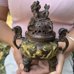 Puede incluir: Un quemador de incienso de bronce con intrincados diseños de dragones y criaturas míticas. El quemador tiene tres patas, asas y una tapa con una pequeña estatua encima. Los detalles se destacan con detalles dorados.