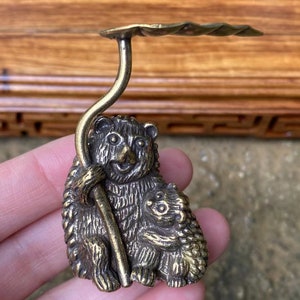 May include: A brass figurine of a bear and cub, holding a curved stem with a leaf-shaped top. The bear is smiling, and the cub is nestled close. The figurine has a detailed, textured surface and a warm, antique finish.
