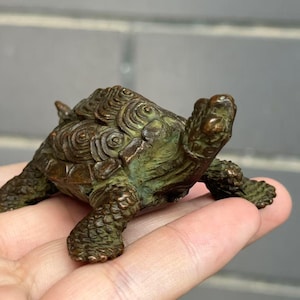 May include: A small, bronze-colored turtle figurine with a detailed shell pattern and textured skin. The figurine is held in a hand, showcasing its size and intricate design. The turtle's head and legs are visible, adding to its realistic appearance.