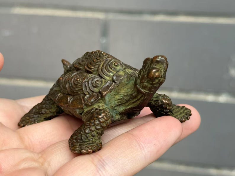 May include: A small, bronze-colored turtle figurine with a detailed shell pattern and textured skin. The figurine is held in a hand, showcasing its size and intricate design. The turtle's head and legs are visible, adding to its realistic appearance.