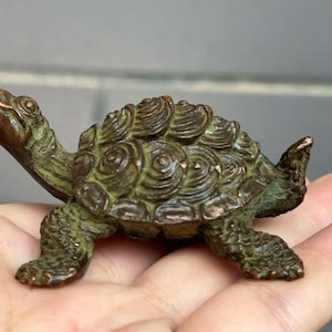 May include: A small, bronze-colored turtle figurine with a detailed shell pattern. The sculpture has a weathered, antique appearance, with green and brown tones. The turtle is posed in a walking position, showcasing its legs and head.