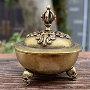May include: A small, antique brass incense burner with a decorative lid and three skull-shaped feet. The lid is adorned with ornate floral patterns and a crown-like finial. The burner has a round bowl shape.
