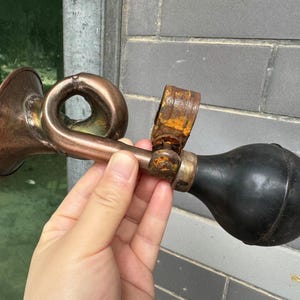 Puede incluir: Un claxon de bicicleta antiguo de cobre y lat&oacute;n con una pera de goma negra. El claxon tiene un dise&ntilde;o curvo y muestra signos de edad y desgaste, con algo de &oacute;xido visible en los componentes met&aacute;licos. El claxon se sostiene en una mano.