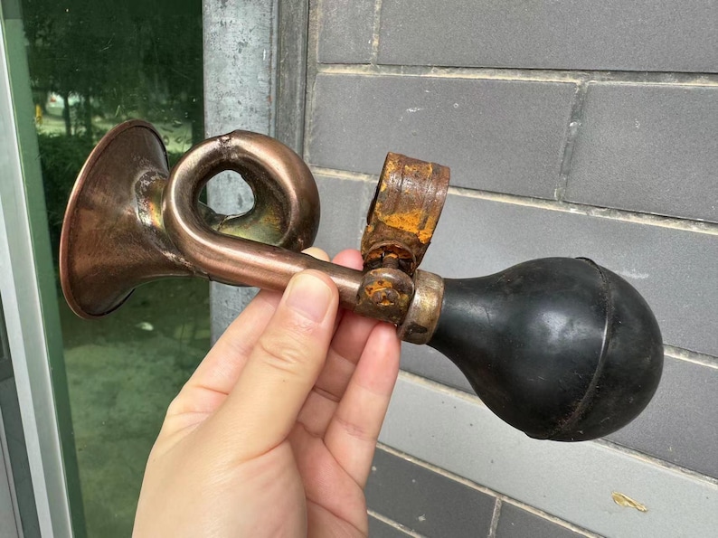 Puede incluir: Un claxon de bicicleta antiguo de cobre y lat&oacute;n con una pera de goma negra. El claxon tiene un dise&ntilde;o curvo y muestra signos de edad y desgaste, con algo de &oacute;xido visible en los componentes met&aacute;licos. El claxon se sostiene en una mano.