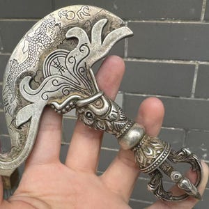 May include: An ornate silver ritual knife with a curved blade. The blade features a detailed dragon design and other decorative elements. The handle has a detailed floral pattern and a ring at the base. The object is held in a hand.