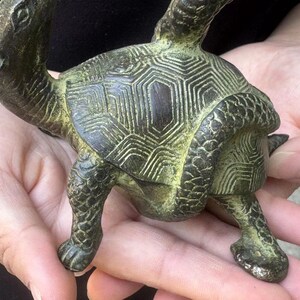 May include: A bronze turtle figurine with a detailed shell pattern and textured skin. The sculpture has a weathered, antique appearance, with green and brown tones. The turtle is depicted in a crawling position, showcasing its legs and head.