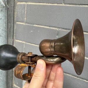 Puede incluir: Un claxon de coche antiguo de lat&oacute;n con una pera de goma negra. El claxon tiene un extremo en forma de campana y una abrazadera para el montaje. El metal tiene un aspecto envejecido, lo que sugiere un uso vintage.