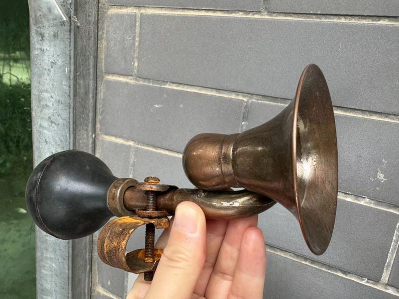 Puede incluir: Un claxon de coche antiguo de lat&oacute;n con una pera de goma negra. El claxon tiene un extremo en forma de campana y una abrazadera para el montaje. El metal tiene un aspecto envejecido, lo que sugiere un uso vintage.