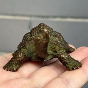 May include: A small, bronze-colored turtle figurine with a detailed shell and textured skin. The sculpture has a weathered, antique appearance, with hints of green patina. The turtle is resting on a hand, showcasing its size and intricate design.
