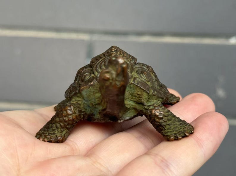 May include: A small, bronze-colored turtle figurine with a detailed shell and textured skin. The sculpture has a weathered, antique appearance, with hints of green patina. The turtle is resting on a hand, showcasing its size and intricate design.