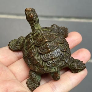 May include: A bronze turtle figurine with a detailed shell pattern and outstretched limbs. The sculpture has a weathered, aged appearance, with a green patina. The turtle is held in a hand, showcasing its size and intricate design.