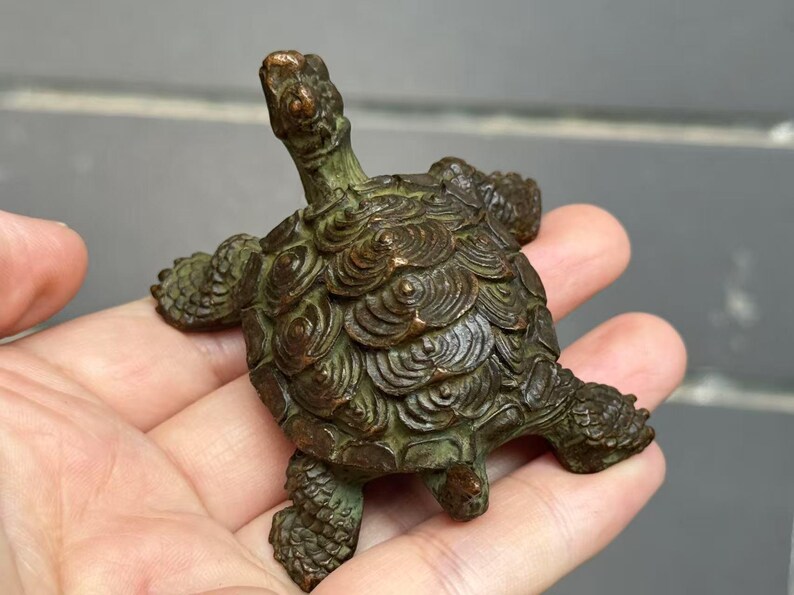 May include: A bronze turtle figurine with a detailed shell pattern and outstretched limbs. The sculpture has a weathered, aged appearance, with a green patina. The turtle is held in a hand, showcasing its size and intricate design.