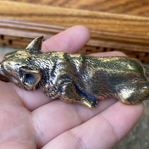 Puede incluir: Escultura de bronce de un perro corgi en posición de descanso. La escultura tiene una superficie texturizada y un tono marrón dorado cálido. Los rasgos del corgi, incluyendo orejas, hocico y patas, están claramente definidos.