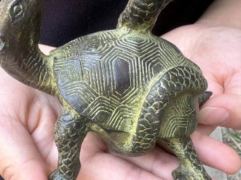 May include: A detailed bronze turtle figurine with a textured shell and intricate patterns. The sculpture has a weathered, antique appearance with green and brown tones. The turtle is held in a person's hands.